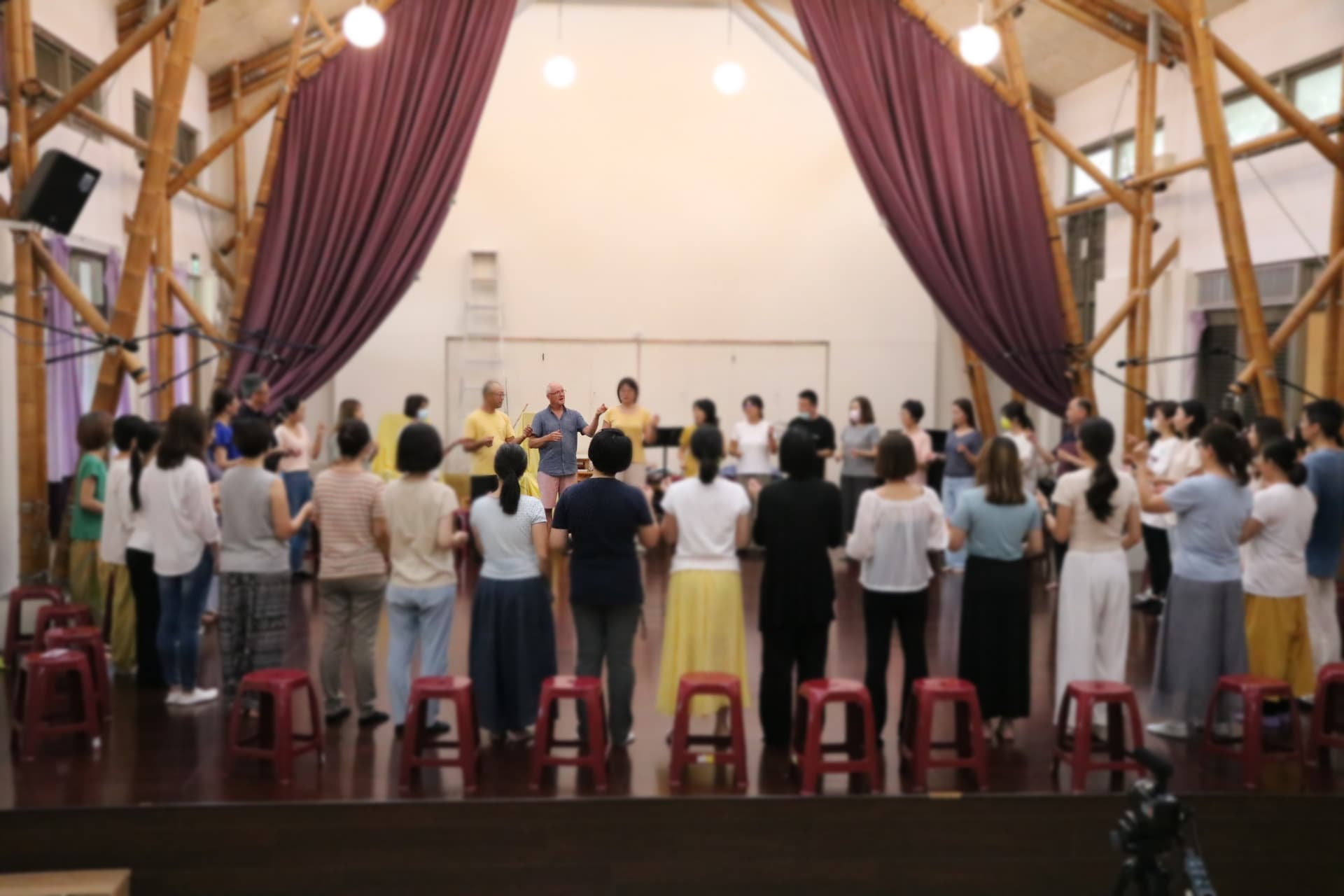 Workshop participants standing in a wide circle, singing together in a bamboo-framed hall